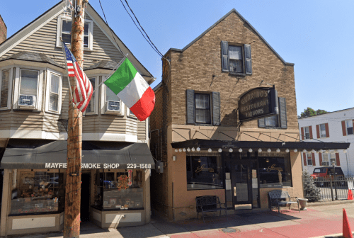 A brick building with a restaurant sign, featuring an Italian flag and an American flag outside, on a sunny day.
