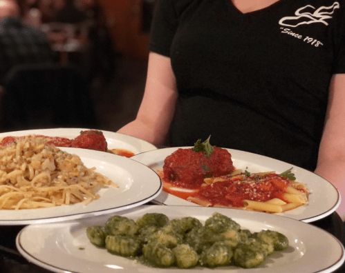 A server holds three plates of pasta dishes, including meatballs, gnocchi, and a creamy pasta, in a restaurant setting.
