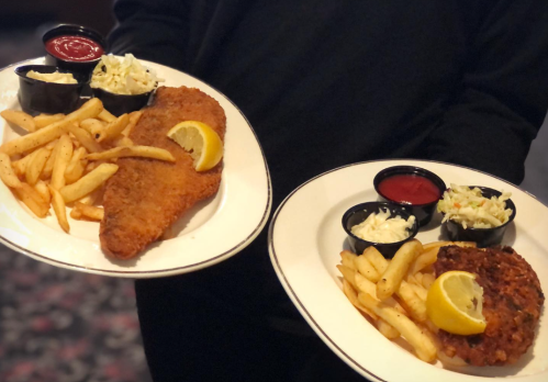 Two plates of fried fish and chicken, served with fries, coleslaw, and dipping sauces, held by a person.