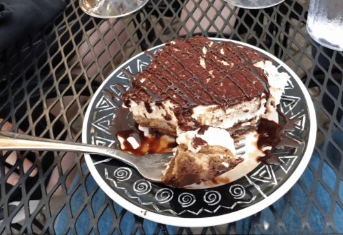 A slice of tiramisu on a decorative plate, drizzled with chocolate sauce, with a fork resting beside it.