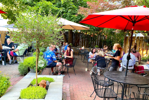 Outdoor dining scene with patrons at tables under red umbrellas, surrounded by greenery and a cozy atmosphere.