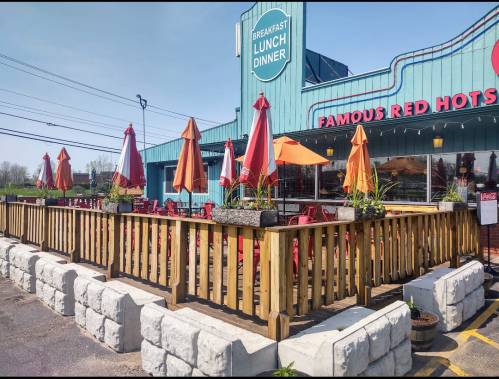 Exterior of a diner with a wooden deck, colorful umbrellas, and a sign reading "Famous Red Hots" in bright colors.