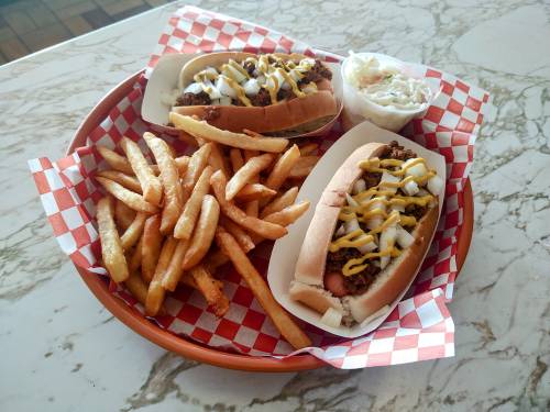 A basket with two chili hot dogs, topped with mustard and onions, served with a side of French fries and coleslaw.