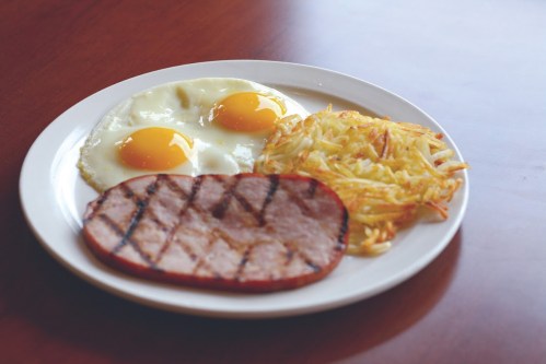 A plate with two fried eggs, grilled ham, and crispy hash browns on a wooden table.