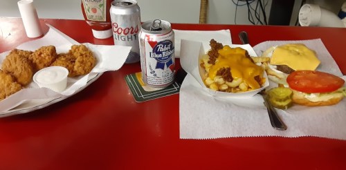 A table with fried chicken, fries topped with cheese, a burger with tomato and pickles, and two cans of beer.