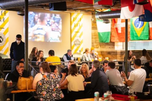 A lively bar scene with people socializing, flags hanging, and a sports game playing on a projector in the background.