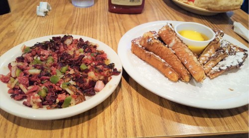 A plate of hash browns with peppers and onions next to a plate of French toast with powdered sugar and a side of syrup.