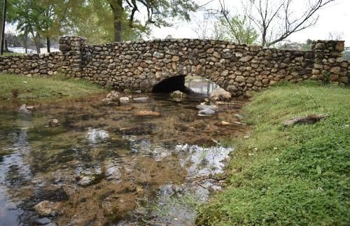 A stone bridge arches over a shallow stream, surrounded by grass and trees.