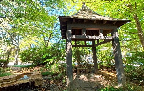A wooden gazebo surrounded by lush greenery and trees, with a stone path and a small water feature nearby.