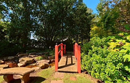 A wooden bridge with red railings crosses a small pond, surrounded by lush greenery and trees under a clear blue sky.
