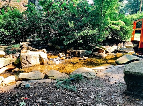 A serene stream flows over rocks, surrounded by lush greenery and a hint of a red bridge in the background.