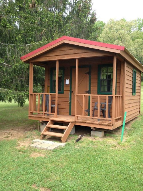 A small wooden cabin with a red roof, porch, and two chairs, surrounded by grass and trees.