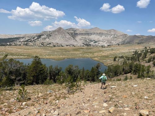 A hiker descends a rocky path towards a serene lake, surrounded by mountains and a clear blue sky.