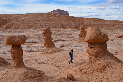 A hiker walks among unique rock formations in a desert landscape under a cloudy sky.