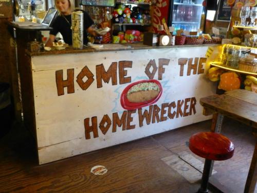 A colorful restaurant counter with a sign reading "Home of the Homewrecker" and a woman serving behind it.