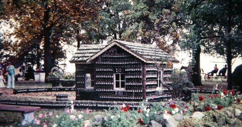 Small house and low garden walls constructed entirely from dark glass bottles in a wooded park.