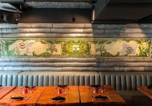 A restaurant interior featuring a large mural of a $5 bill on a textured wall above wooden tables and chairs.