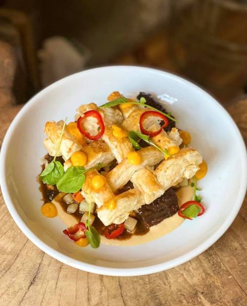 A plate of flaky pastry topped with beef, colorful sauces, and fresh herbs, served on a rustic wooden surface.