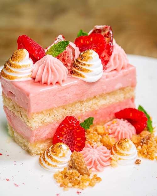 A slice of pink cake layered with cream, topped with strawberries, meringue, and decorative swirls on a white plate.