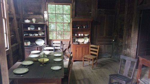 A rustic room with a wooden table set for a meal, shelves of dishes, and a chair in a cozy, dimly lit space.