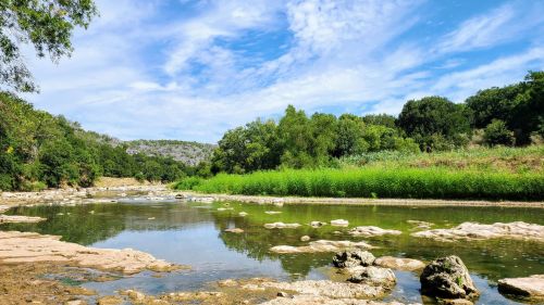 A serene river scene with clear water, rocky banks, and lush greenery under a blue sky with scattered clouds.