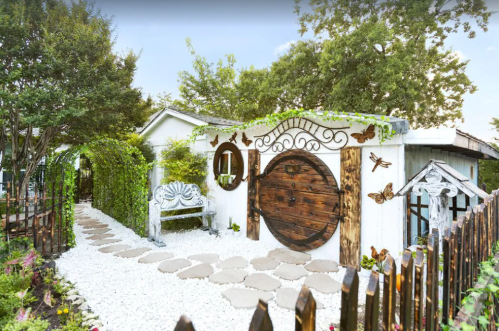 Charming garden entrance with a wooden door, stone path, and lush greenery, featuring decorative elements and a bench.