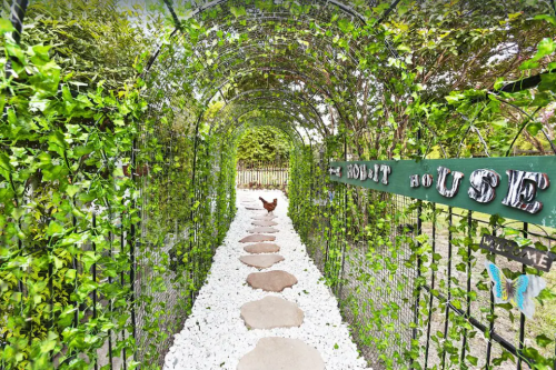 A stone path leads through a green archway covered in vines, with a sign reading "Hobbit House" at the entrance.
