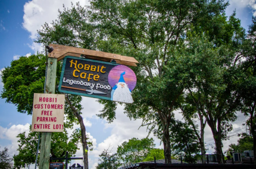 Sign for Hobbit Cafe featuring a wizard graphic, surrounded by trees and a clear blue sky. Free parking for customers.