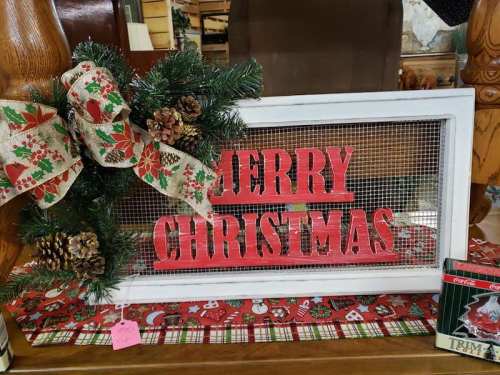 A festive sign reading "Merry Christmas" adorned with greenery, pinecones, and a decorative bow.