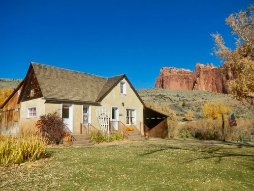 A quaint house with a sloped roof, surrounded by greenery, set against a backdrop of red rock formations and a clear blue sky.