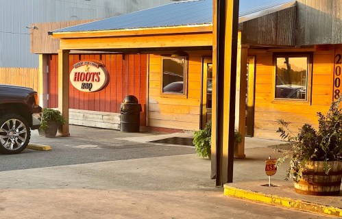Exterior of Hoot's BBQ restaurant, featuring wooden walls, a sign, and a parking area in the foreground.