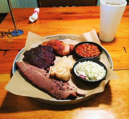 A platter of barbecue featuring brisket, chicken, baked beans, coleslaw, and a slice of bread on a wooden table.