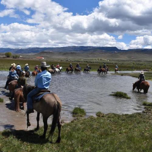 A group of horseback riders crossing a shallow pond in a scenic landscape with mountains and clouds in the background.