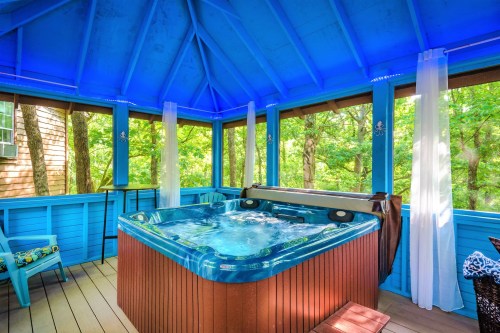 A bright blue gazebo with a hot tub surrounded by trees, featuring sheer curtains and a cozy seating area.