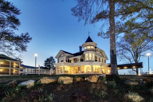 A large, elegant house with a turret, surrounded by trees, illuminated at dusk.