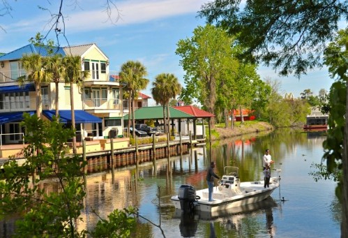 A boat with two people floats on a calm waterway, surrounded by trees and colorful buildings along the shore.