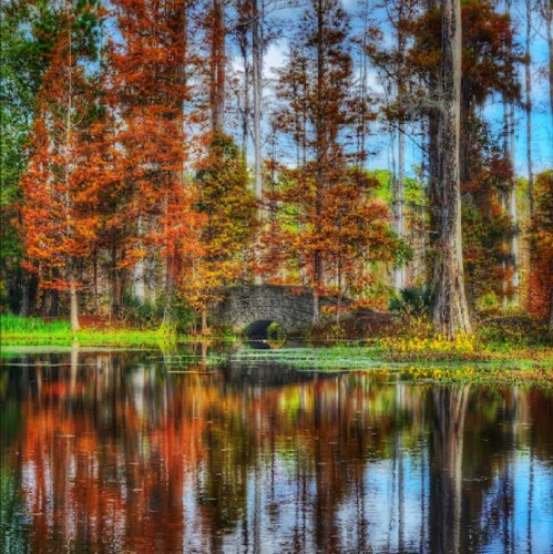 A serene pond reflects vibrant autumn trees and a stone bridge under a clear blue sky.