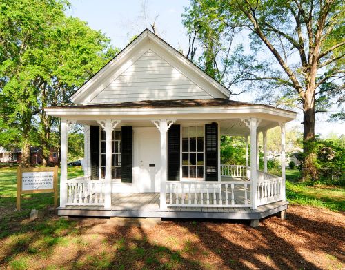 A small white house with a porch, surrounded by trees and greenery, featuring black shutters and a sign in front.