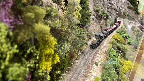 A model train travels along a scenic track surrounded by lush greenery and rocky terrain.