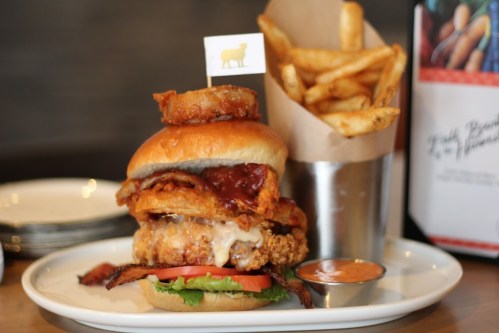 A stacked burger with fried chicken, bacon, lettuce, and tomato, served with crispy fries in a cup.