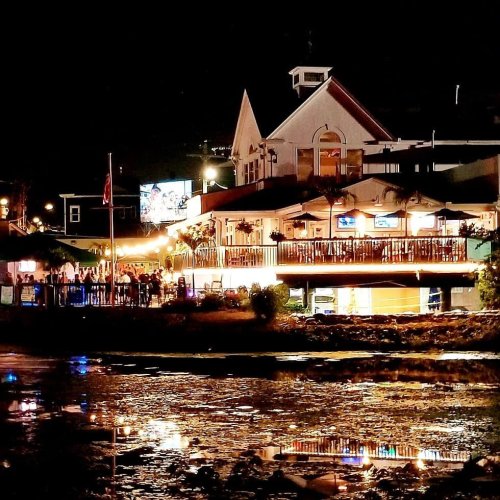 A brightly lit restaurant by the water at night, reflecting lights on the calm surface.