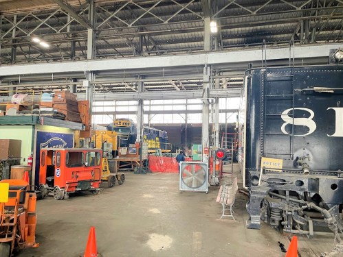 Interior of a train workshop with various vehicles, tools, and equipment, featuring a large black train engine.