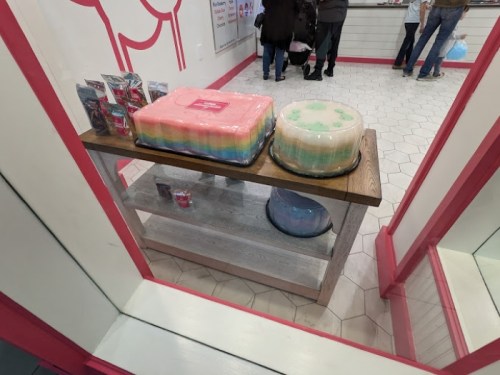 Two colorful layered cakes on a wooden table inside a shop, with customers in the background.