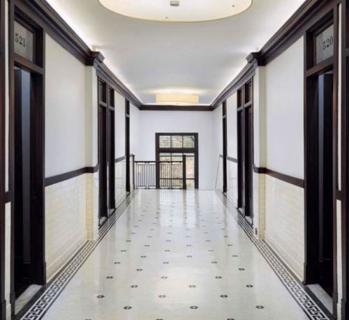 A long, well-lit hallway with dark wooden doors and a patterned tile floor, featuring room numbers on the doors.