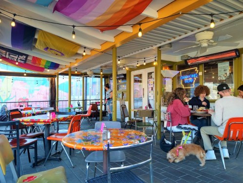 A vibrant café patio with colorful decor, tables, and patrons enjoying their time, including a dog on a leash.