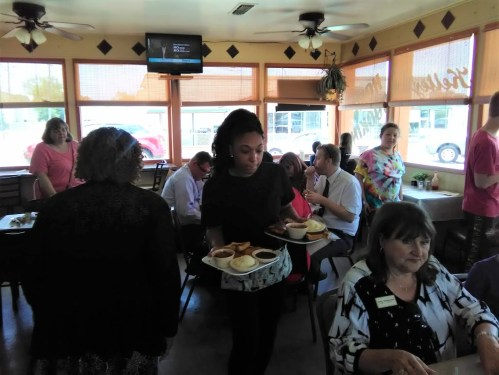 A busy restaurant scene with a server carrying trays of food, while patrons chat at tables.