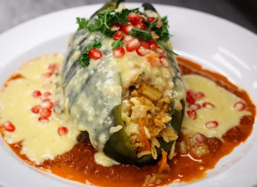 A stuffed green pepper topped with pomegranate seeds and drizzled with creamy sauce, served on a plate with red sauce.