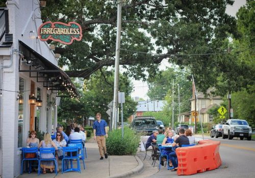Outdoor dining scene at a restaurant with people sitting at blue tables, surrounded by trees and street activity.