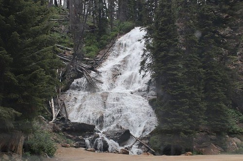 A cascading waterfall flows down rocky terrain, surrounded by lush green trees and a forested landscape.