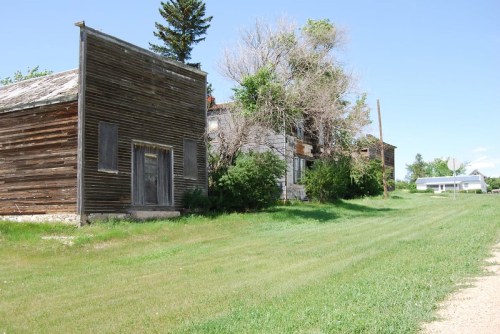 Abandoned buildings line a grassy road under a clear blue sky, with trees and a distant structure visible.